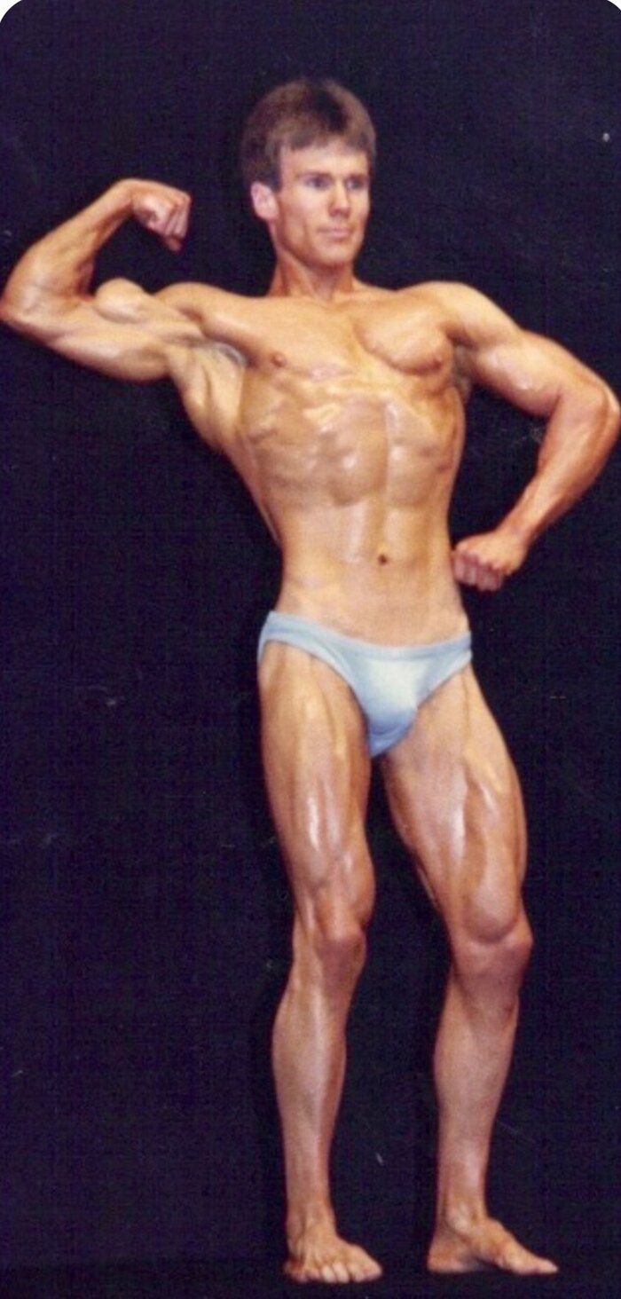 Dr. David Waring competing on stage as an NPC bodybuilder in a front double-biceps pose during his Mr. Michigan championship years